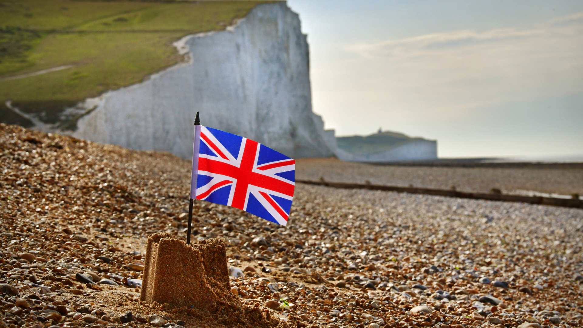 British Union Jack flag sticking out of the top of small sandcastle on pebbled each with cliffs in background
