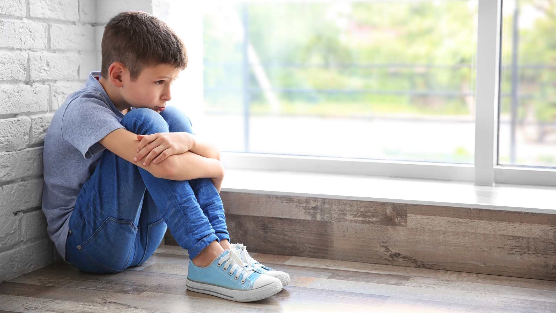 Sad boy with his knees tucker up under his chin by a window
