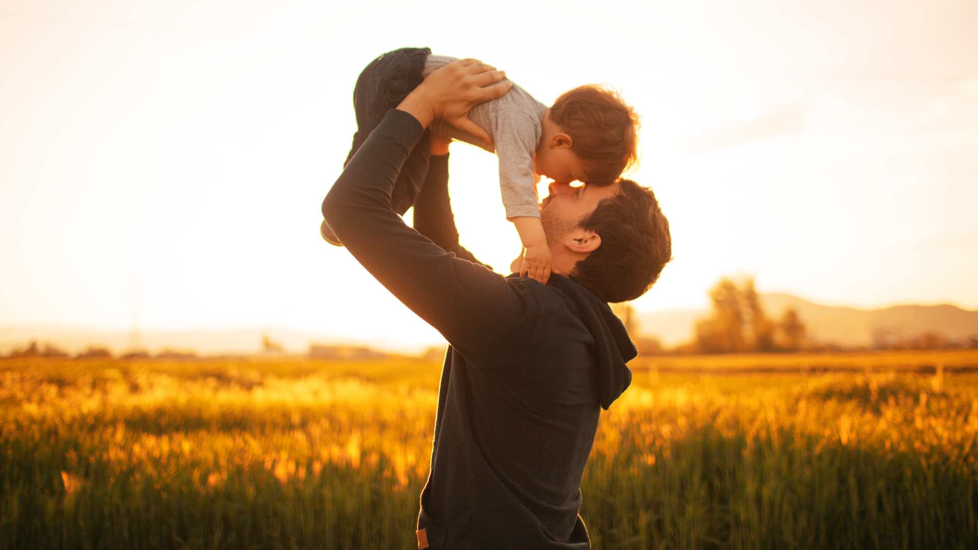 A father lovingly holding his son in their air and touching noses with each other in sunset field
