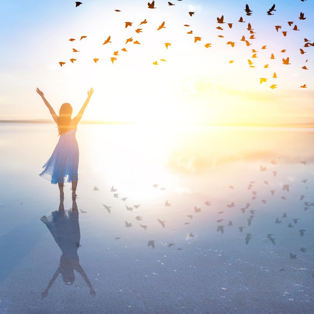 Woman in summer dress on beach with arms in the air with flocks of birds flying around her