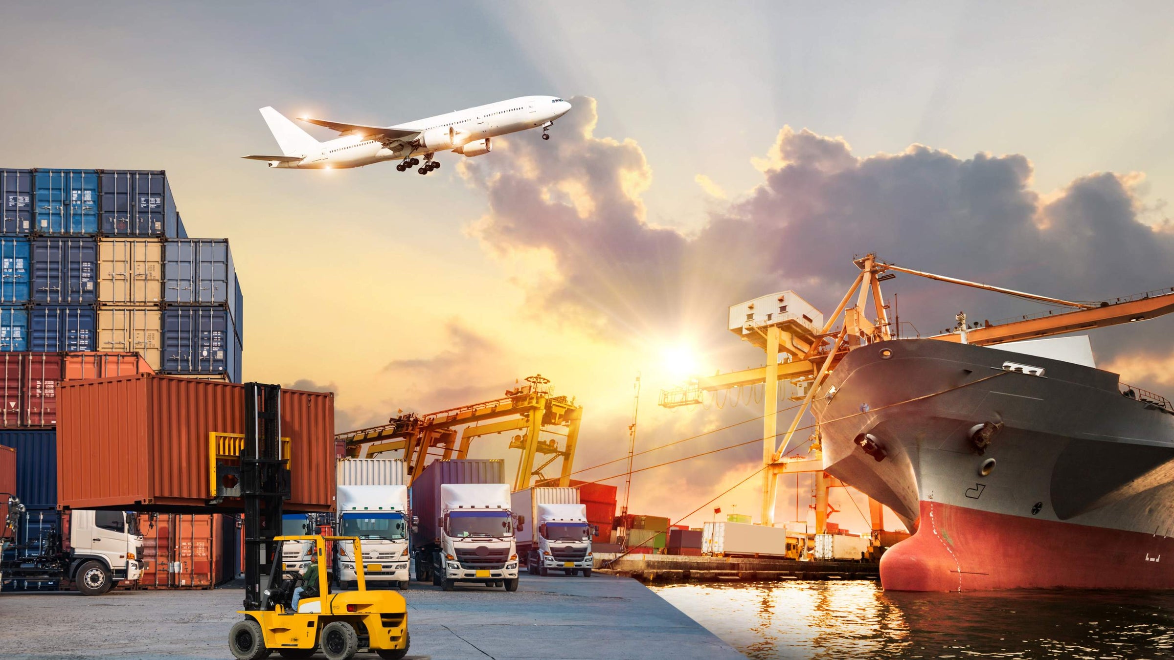 Cargo containers at a dock with a large ship, lorries and an aircraft flying overhead